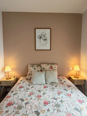 A bedroom with a floral bedspread, two bedside lamps, a framed artwork on the wall, and a light switch. - Suite Adélaïde, Élégance Rétro au cœur de la ville (Landerneau)