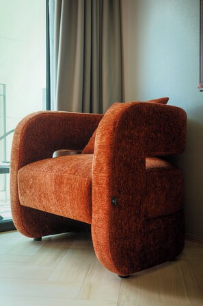 Ein moderner, orangefarbener Sessel mit ungewöhnlichem Design, platziert in einem Raum mit Holzboden und einem großen Fenster mit Vorhängen.