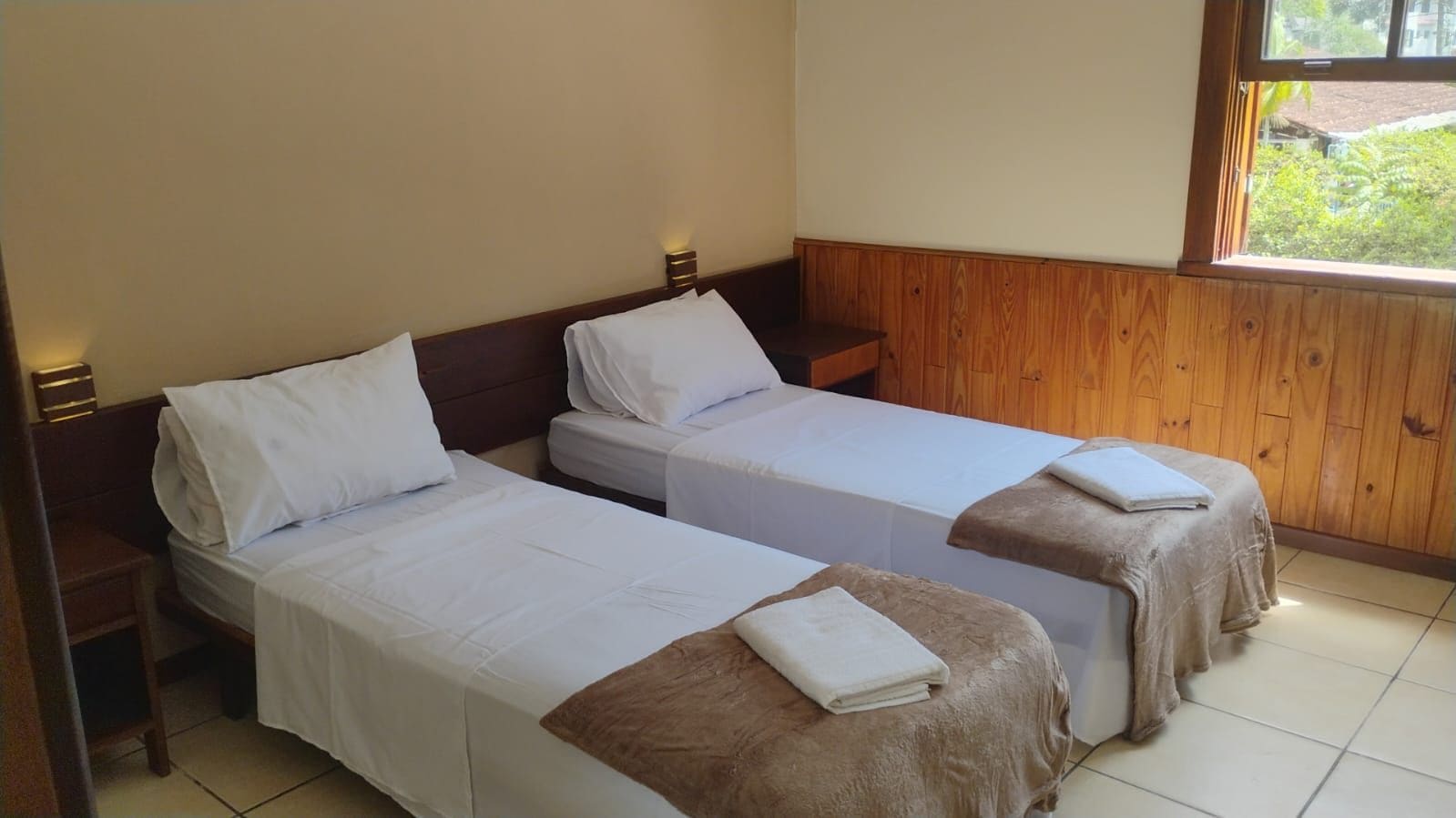 A room with two single beds, wooden headboards, a small table, and a window with a view of greenery.