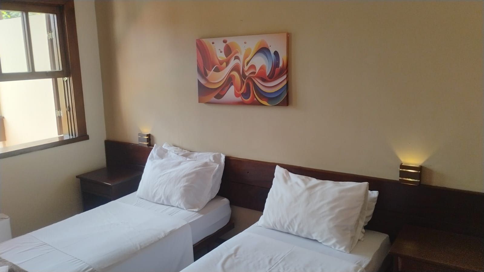 Habitación de hotel con dos camas individuales, una mesita de noche con lámpara y un cuadro abstracto de colores en la pared.