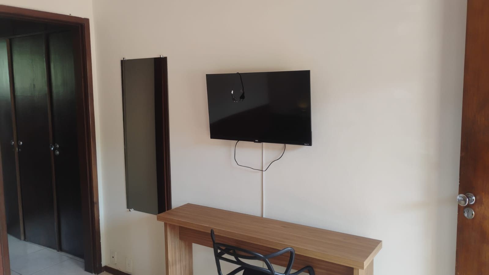 Une pièce avec un bureau en bois, une chaise, un téléviseur à écran plat fixé au mur et une porte dotée d’un cadre foncé.