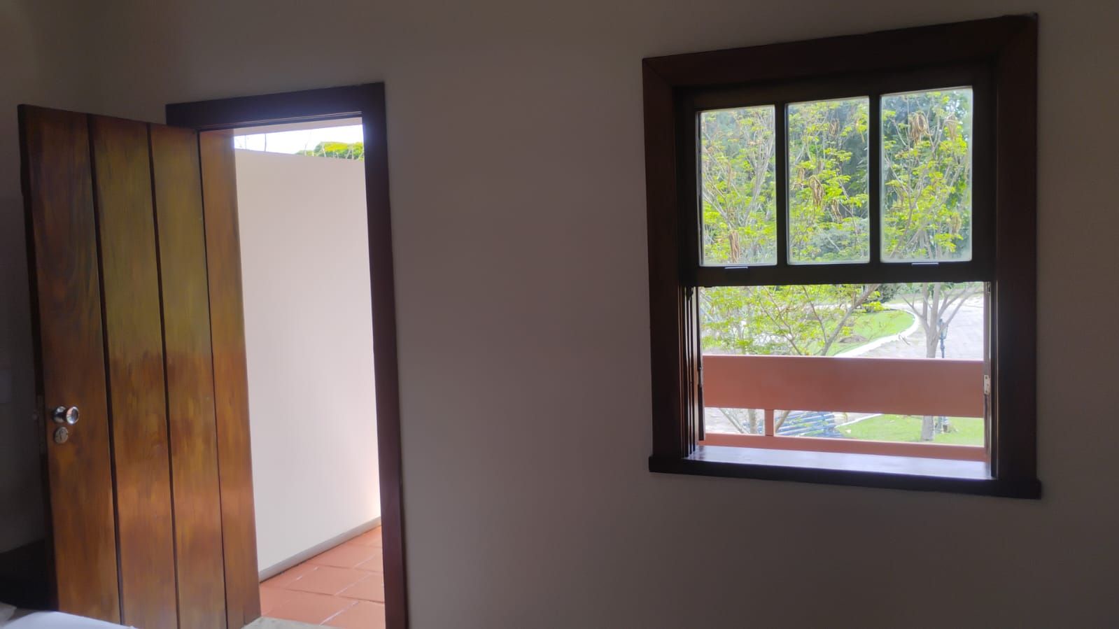 A room with a wooden door, a window with a view of greenery, and a balcony.