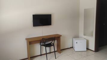 A room with a wooden desk, a black chair, a television mounted on the wall, and a small white appliance on the floor.