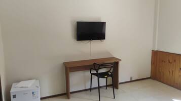 A room with a wooden desk, a black chair, a television mounted on the wall, and a white appliance on the floor.