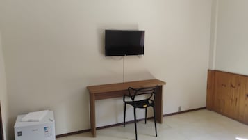 A room with a wooden desk, a black chair, a television mounted on the wall, and a white appliance on the floor.