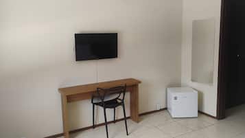 Una habitación con un escritorio de madera, una silla negra, un televisor fijado a la pared y un pequeño electrodoméstico blanco en el suelo.