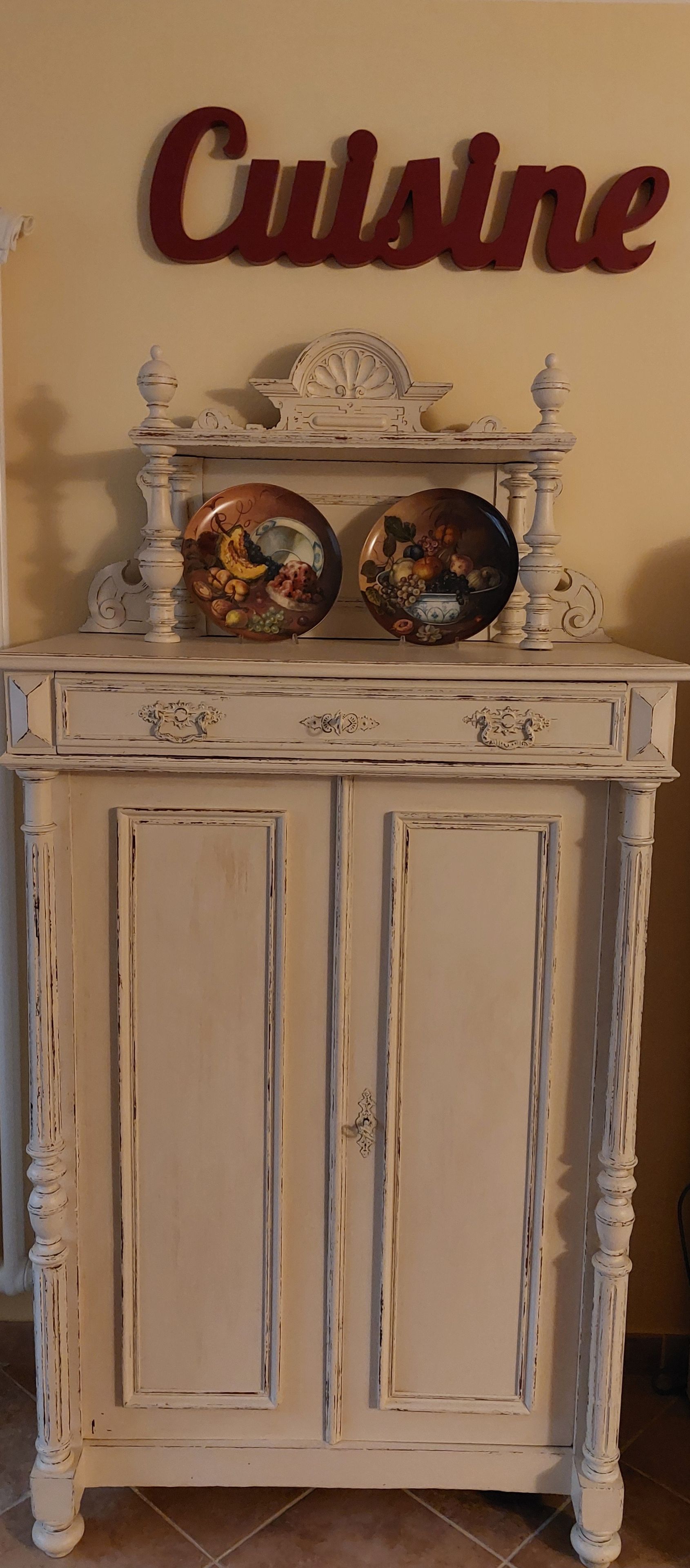 A white cabinet with two decorative plates on top, a wall sign reading 'Cuisine,' and a door with a handle.