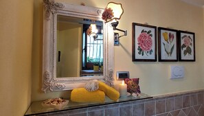 Un baño con un espejo grande y decorativo, una lámpara de pared y grabados botánicos enmarcados.