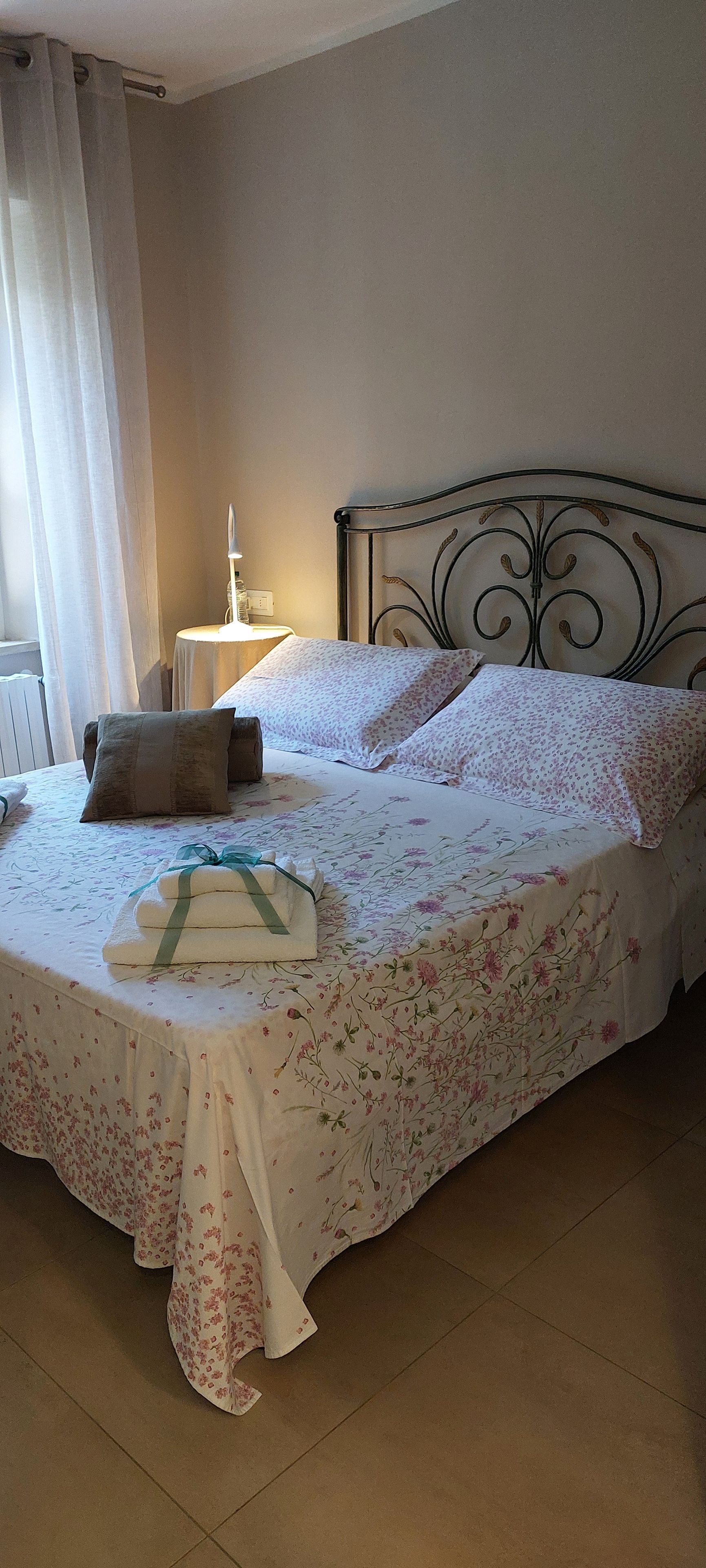 Un letto rifatto con lenzuola a motivi floreali, un comodino con una lampada e una finestra con tende.