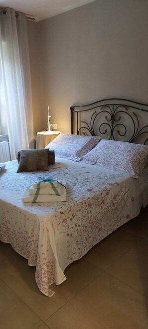 Un letto rifatto con lenzuola a motivi floreali, un comodino con una lampada e una finestra con tende.