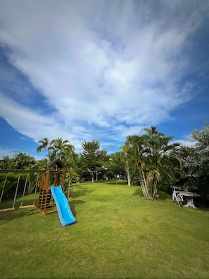 A grassy backyard with a wooden play structure, including a slide, and a bicycle. - Private 3-Bed Villa · Huge 860m² Garden · Mountain View · Gated · Long Stays (Tambon Nong Kae)