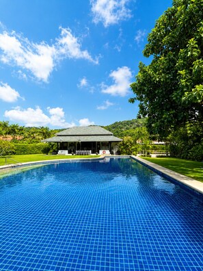 A swimming pool with a tiled surface, surrounded by a grassy area and a building with a roof. - Private 3-Bed Villa · Huge 860m² Garden · Mountain View · Gated · Long Stays (Tambon Nong Kae)