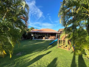 Uma casa com varanda coberta, escorrega e gramado cercado por palmeiras.