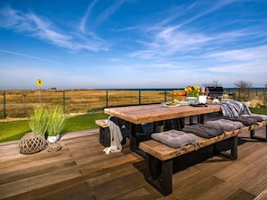 An outdoor dining area with a wooden table and benches, overlooking a fenced field and the sea.