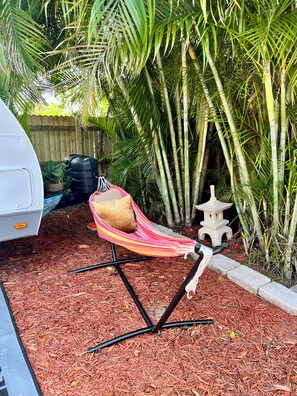 A hammock chair with a cushion, a small stone lantern, and a white vehicle in a garden with bamboo and palm trees. - Cozy 1-bedroom mobile home in soothing Miami with AC (Miami)