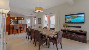Un coin repas avec une table et des chaises en bois, une cuisine à l’arrière-plan et un téléviseur fixé au mur.