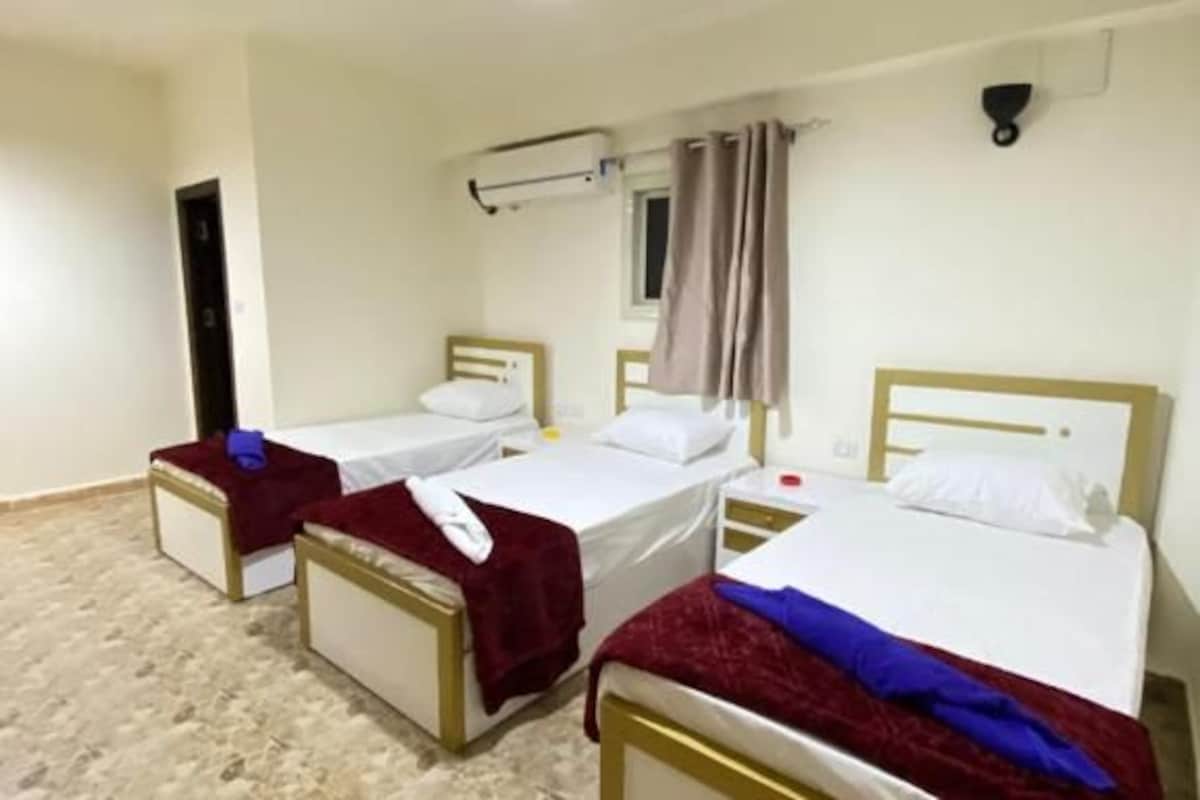 Una camera d'albergo con tre letti singoli, una lampada a soffitto, una finestra con tende e un condizionatore d'aria a parete.