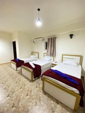 Una camera d'albergo con tre letti singoli, una lampada a soffitto, una finestra con tende e un condizionatore d'aria a parete.