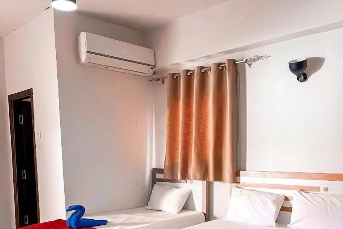 Una camera d'albergo con due letti, una coperta rossa e una bianca, un plaid rosso sul letto bianco, una testiera di legno, un condizionatore d'aria a muro e una tenda.