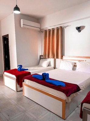 Una camera d'albergo con due letti, una coperta rossa e una bianca, un plaid rosso sul letto bianco, una testiera di legno, un condizionatore d'aria a muro e una tenda.