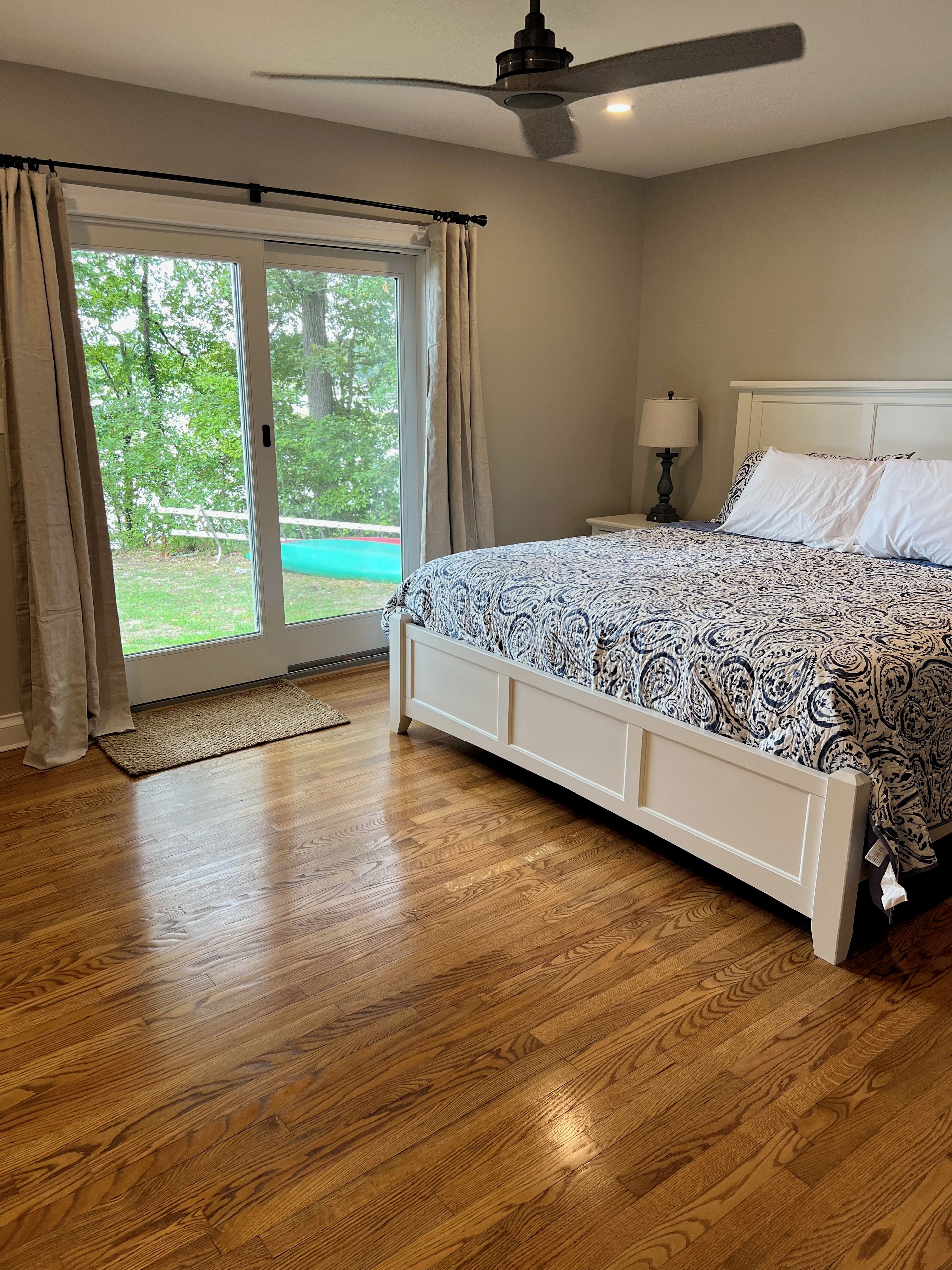 A bedroom with a large bed, a ceiling fan, and a view of a green outdoor area.
