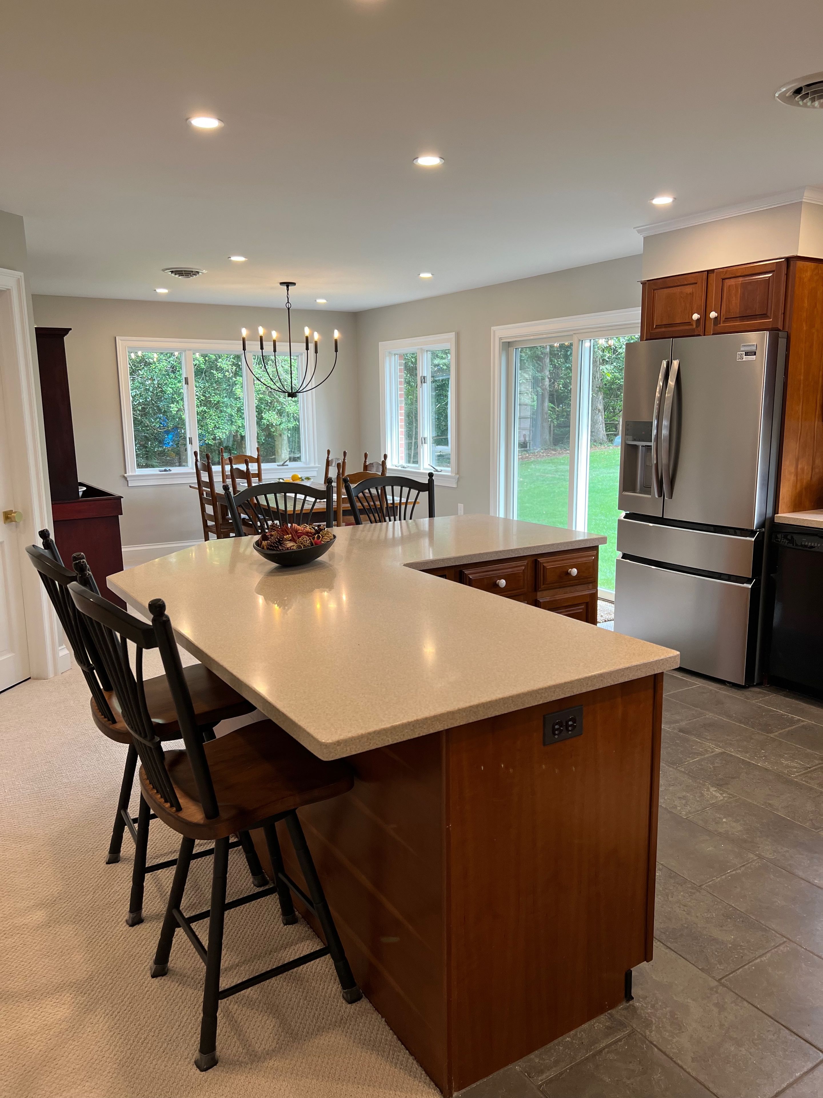 A modern kitchen with a central island, wooden chairs, a stainless steel refrigerator, and large windows.