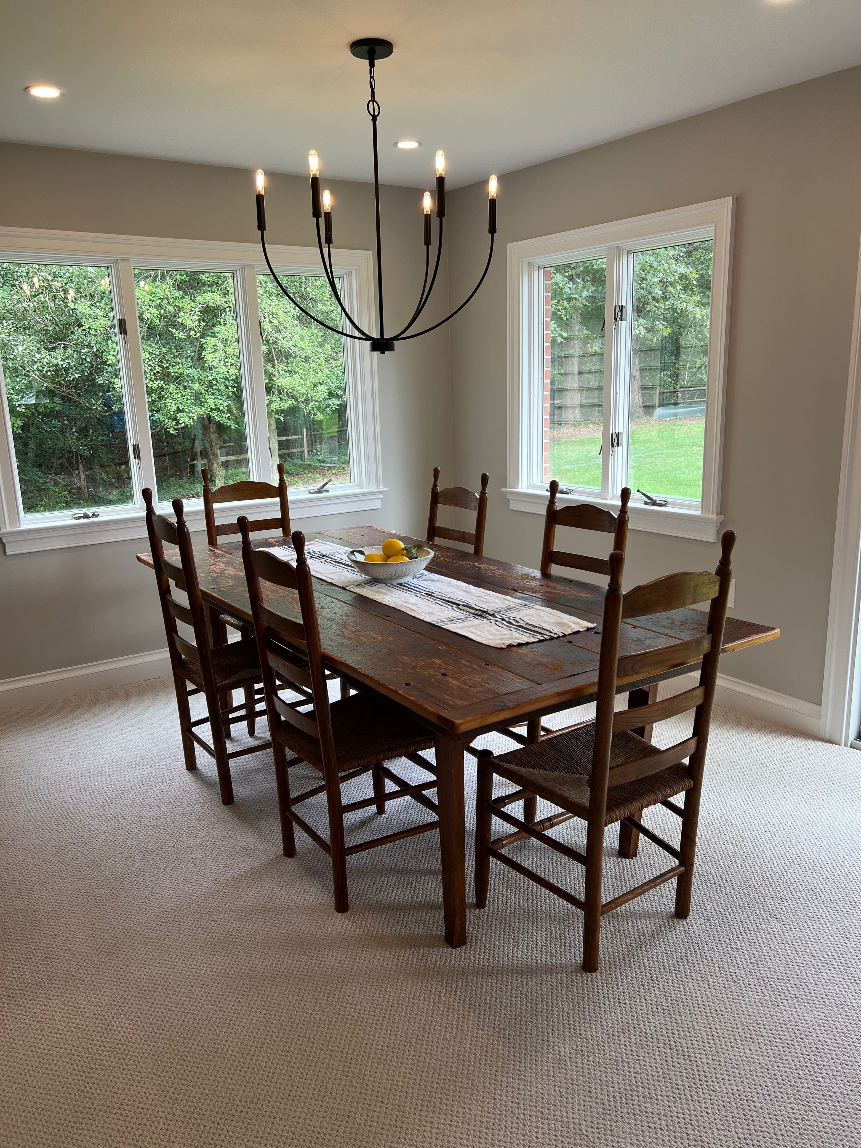 A dining room with a large wooden table, six chairs, a chandelier, and large windows with a view of greenery.