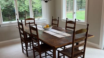 A dining room with a large wooden table, six chairs, a chandelier, and large windows with a view of greenery.