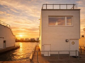 Ein Gebäude am Wasser mit einem Steg, einem Segelboot im Fenster und einem Sonnenuntergang im Hintergrund.