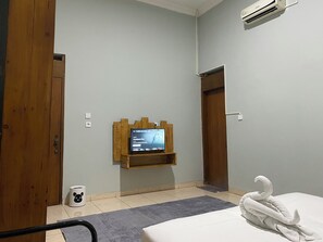 A room with a bed, a mounted TV, a wooden shelf, a white trash bin, and a door.