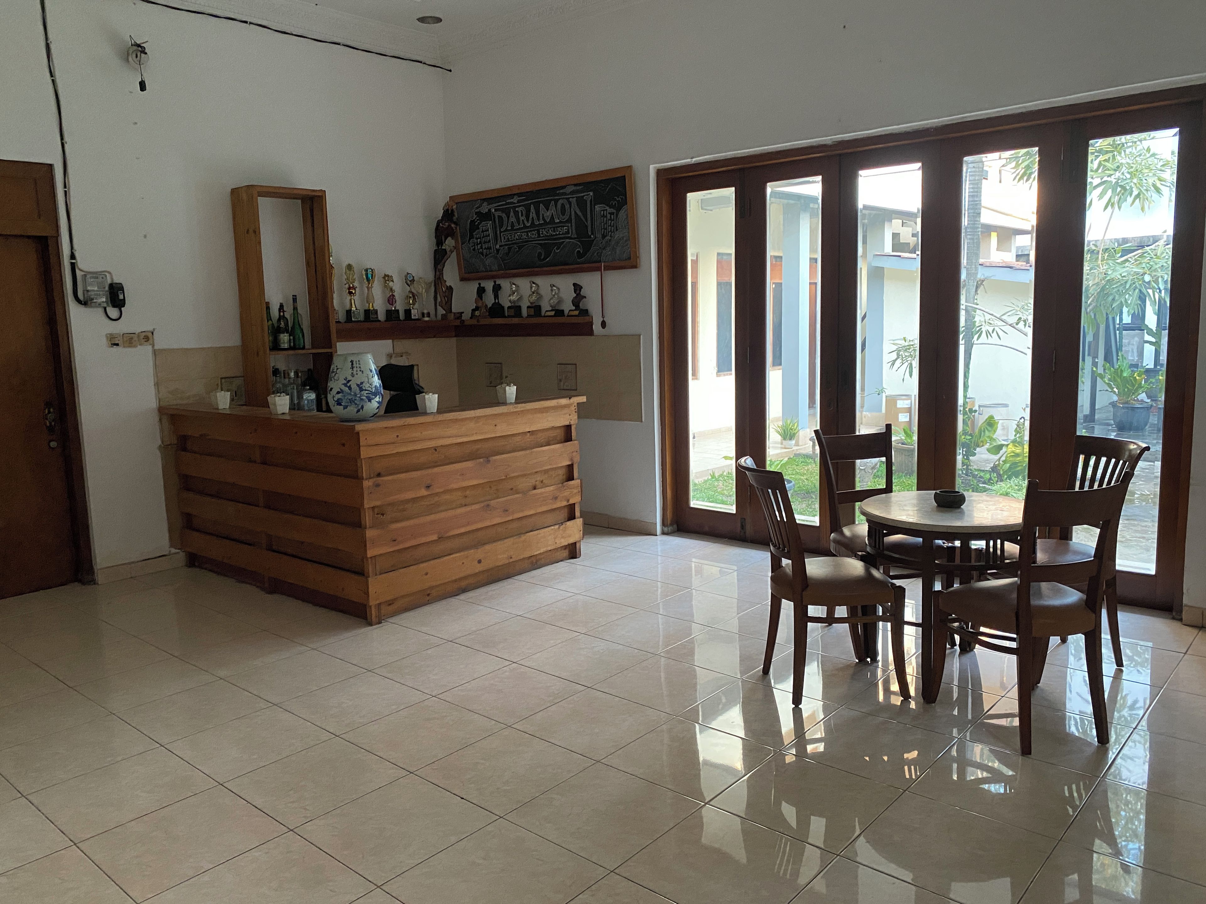 Un área de recepción con mostrador de madera, un espejo y un letrero en una pizarra. Hay un comedor con mesa y sillas, y vistas al exterior a través de amplias ventanas.
