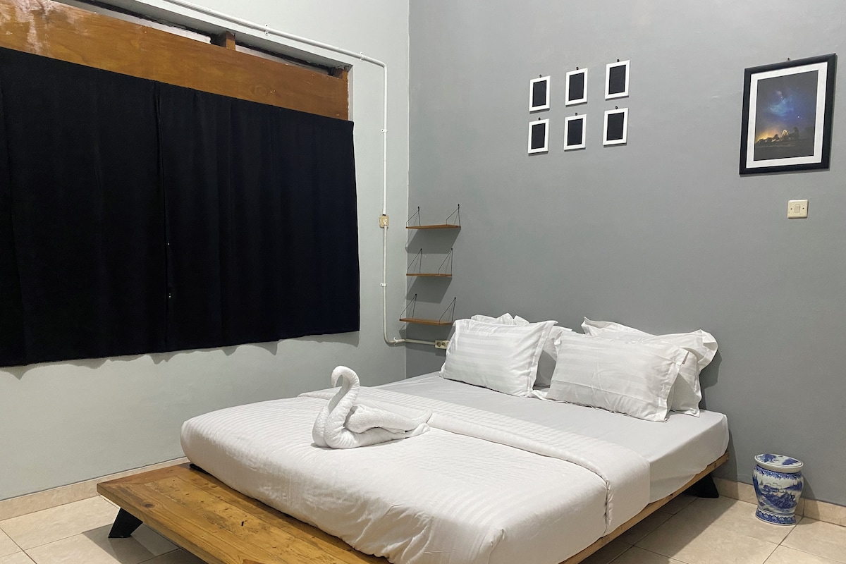 A bedroom with a bed, a wooden platform, a black curtain, a white wall with picture frames, and a small blue vase.