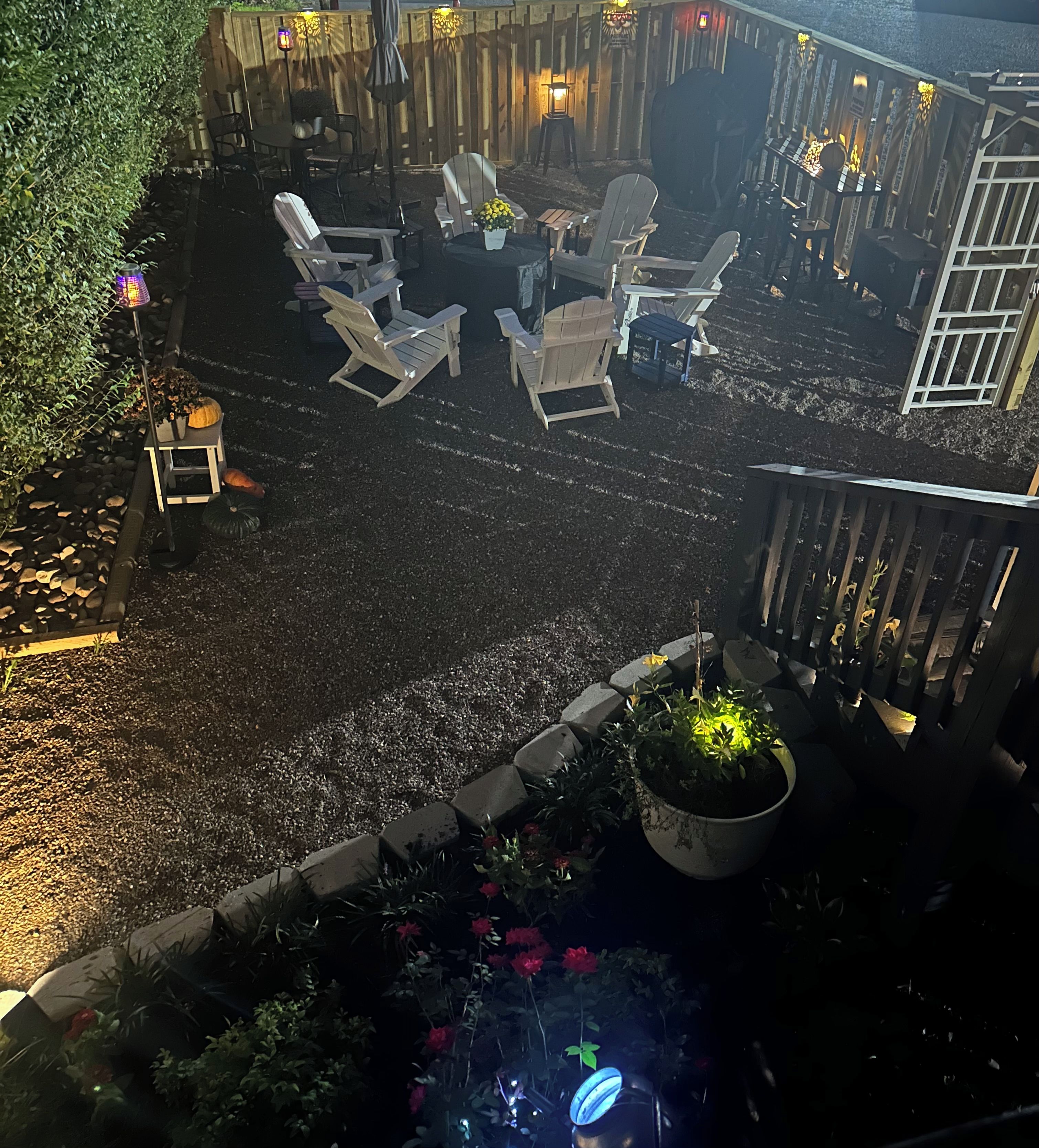 An outdoor seating area with Adirondack chairs and a table, surrounded by a wooden fence and potted plants.