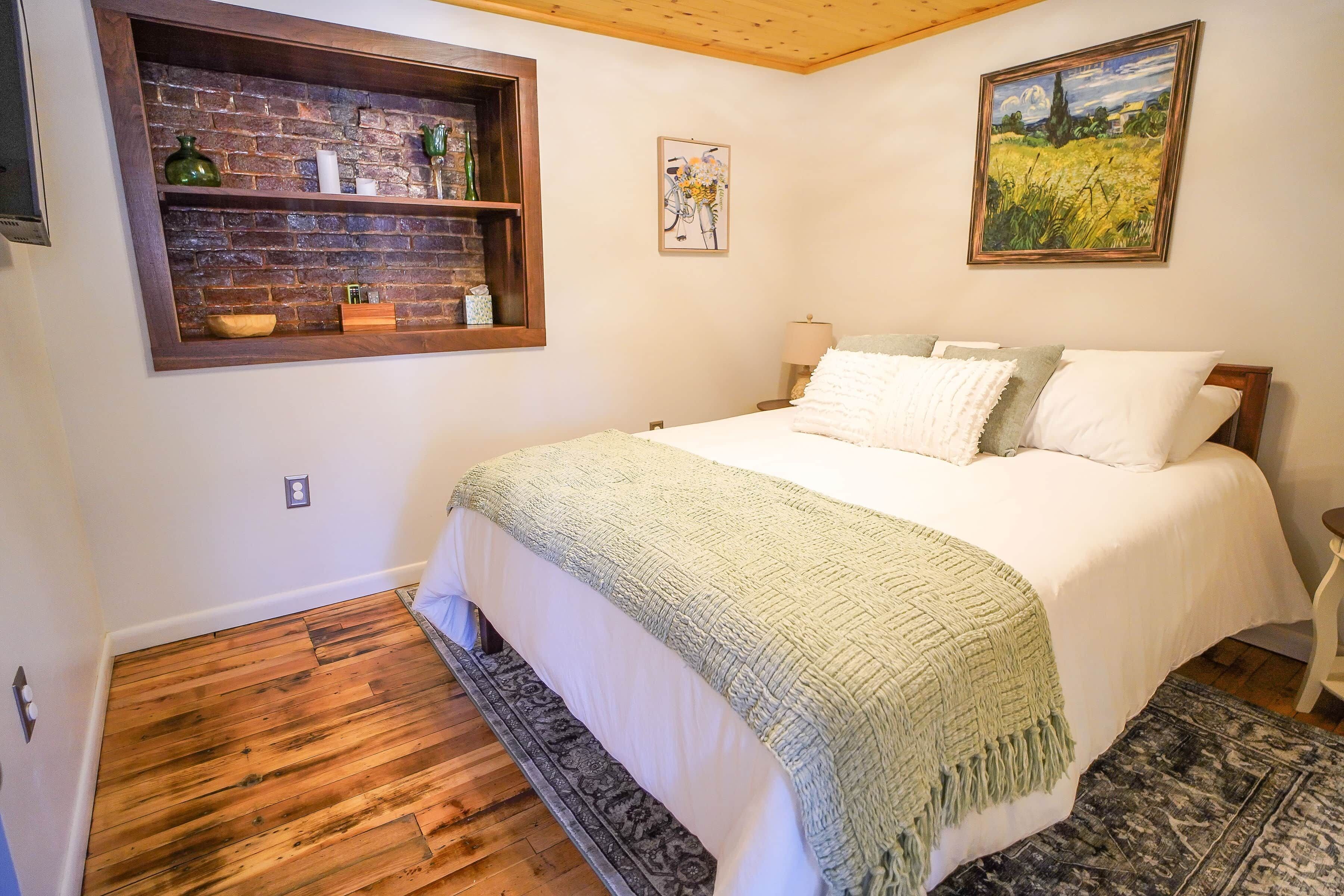 A bedroom with a wooden floor, a bed with white bedding, a green throw blanket, a brick wall shelf with decorative items, and a painting above the bed.