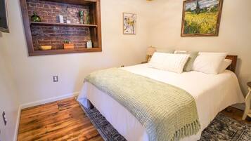 A bedroom with a wooden floor, a bed with white bedding, a green throw blanket, a brick wall shelf with decorative items, and a painting above the bed.