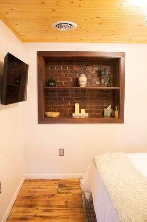 A room with a wooden ceiling, a brick wall shelf with decorative items, a bed with a white blanket, and a wooden floor.