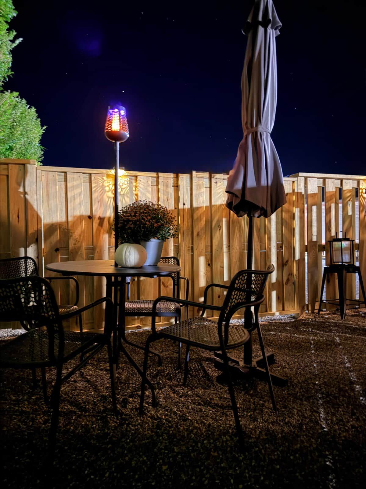 An outdoor patio area with a round table, four chairs, a potted plant, and a hanging umbrella.