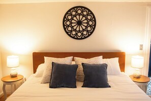 Un lit bien fait, avec du linge de lit blanc et plusieurs oreillers, deux lampes de chevet et un élément décoratif mural au-dessus de la tête de lit.