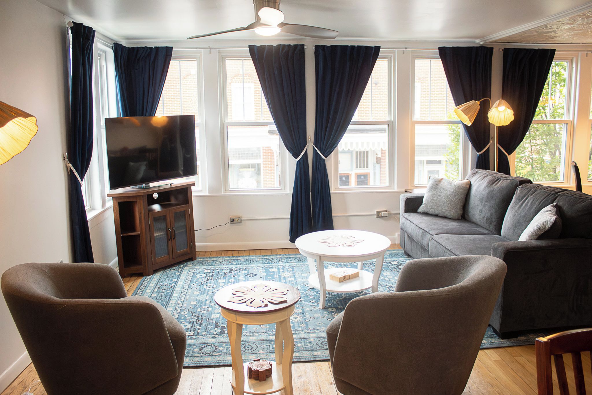 A living room with a sofa, chairs, a TV, a ceiling fan, and a view of buildings through large windows.