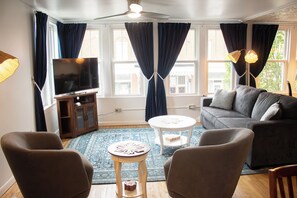 A living room with a sofa, chairs, a TV, a ceiling fan, and a view of buildings through large windows.