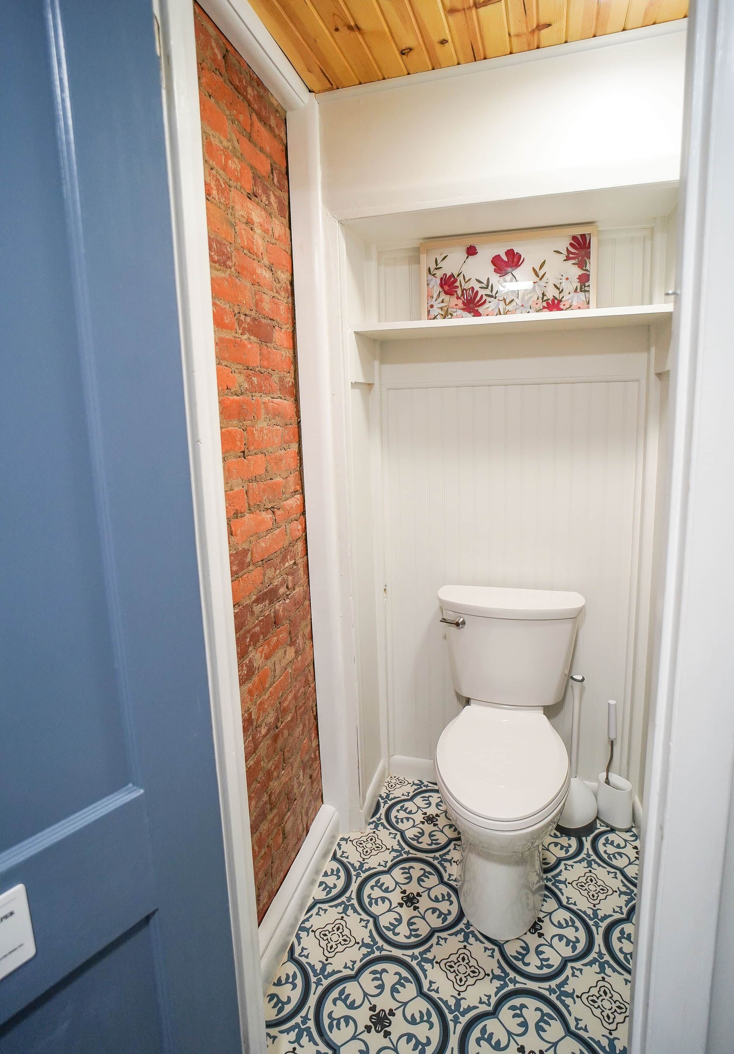 A small bathroom with a toilet, a white shelf with a floral decoration, and a patterned floor.