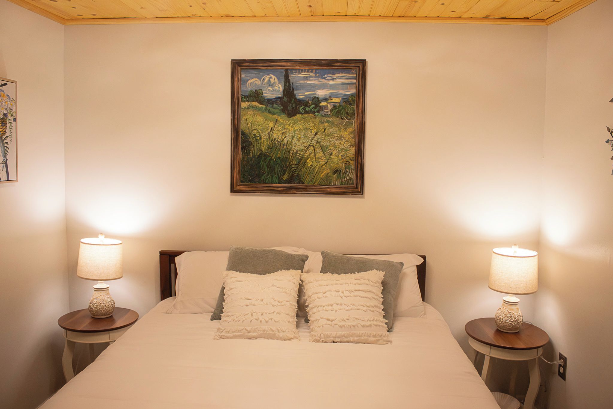 A bedroom with a bed, two bedside lamps, a painting on the wall, and a small table with a lamp.