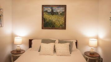 A bedroom with a bed, two bedside lamps, a painting on the wall, and a small table with a lamp.