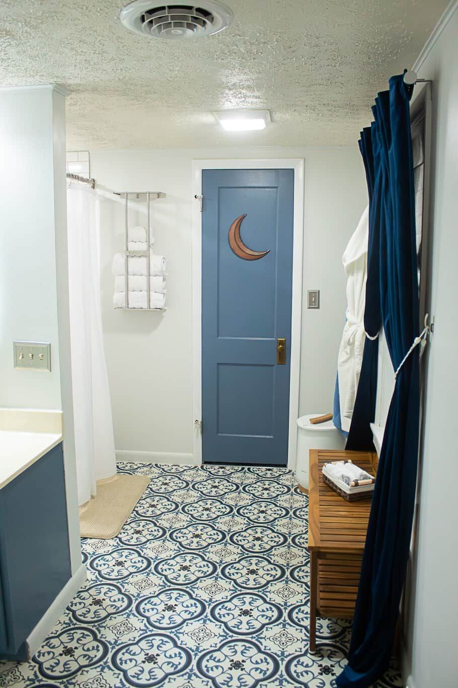 Un baño con piso estampado, una puerta azul con forma de luna creciente, un perchero y un banco de madera.