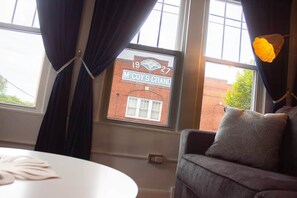 A room with a view of a brick building through a window, a round white table, and a grey sofa with a patterned cushion.