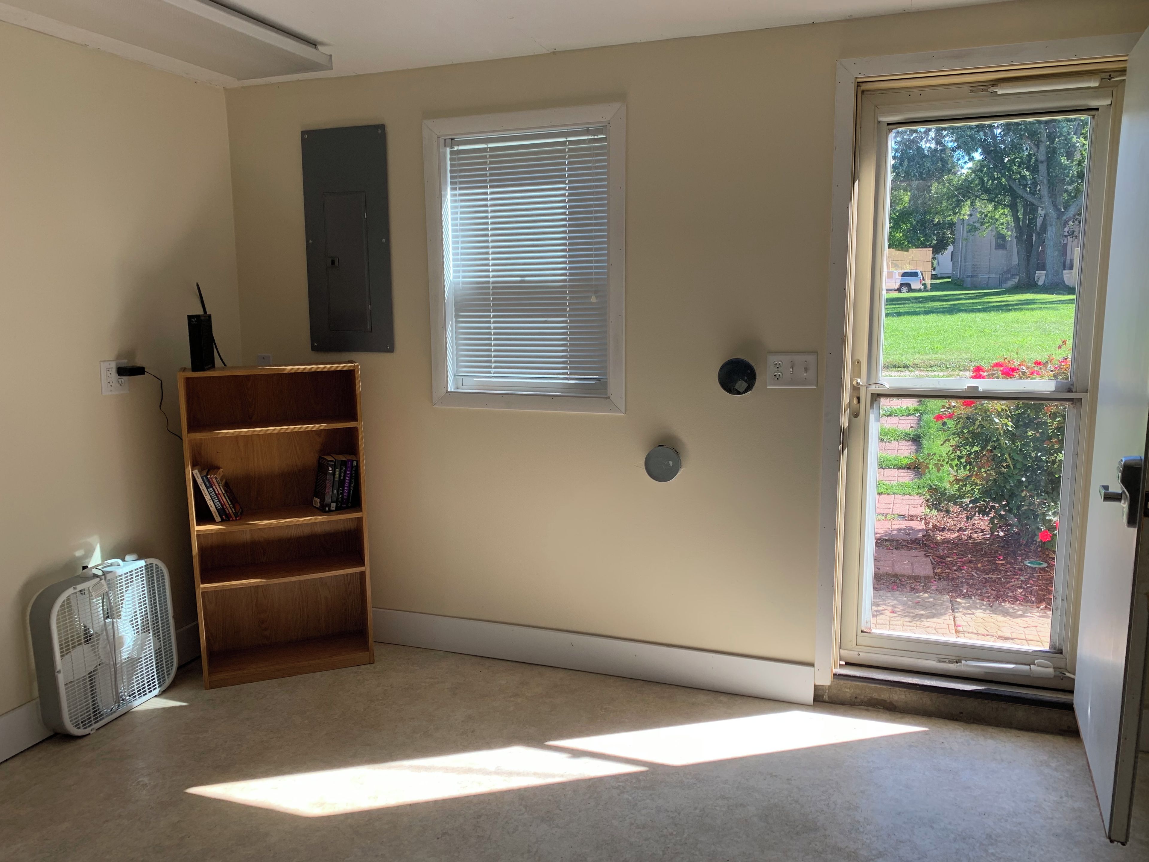 Ein Zimmer mit einer Tür zu einem Garten, einem Bücherregal, einem an der Wand montierten Stromkasten und einem Standventilator.