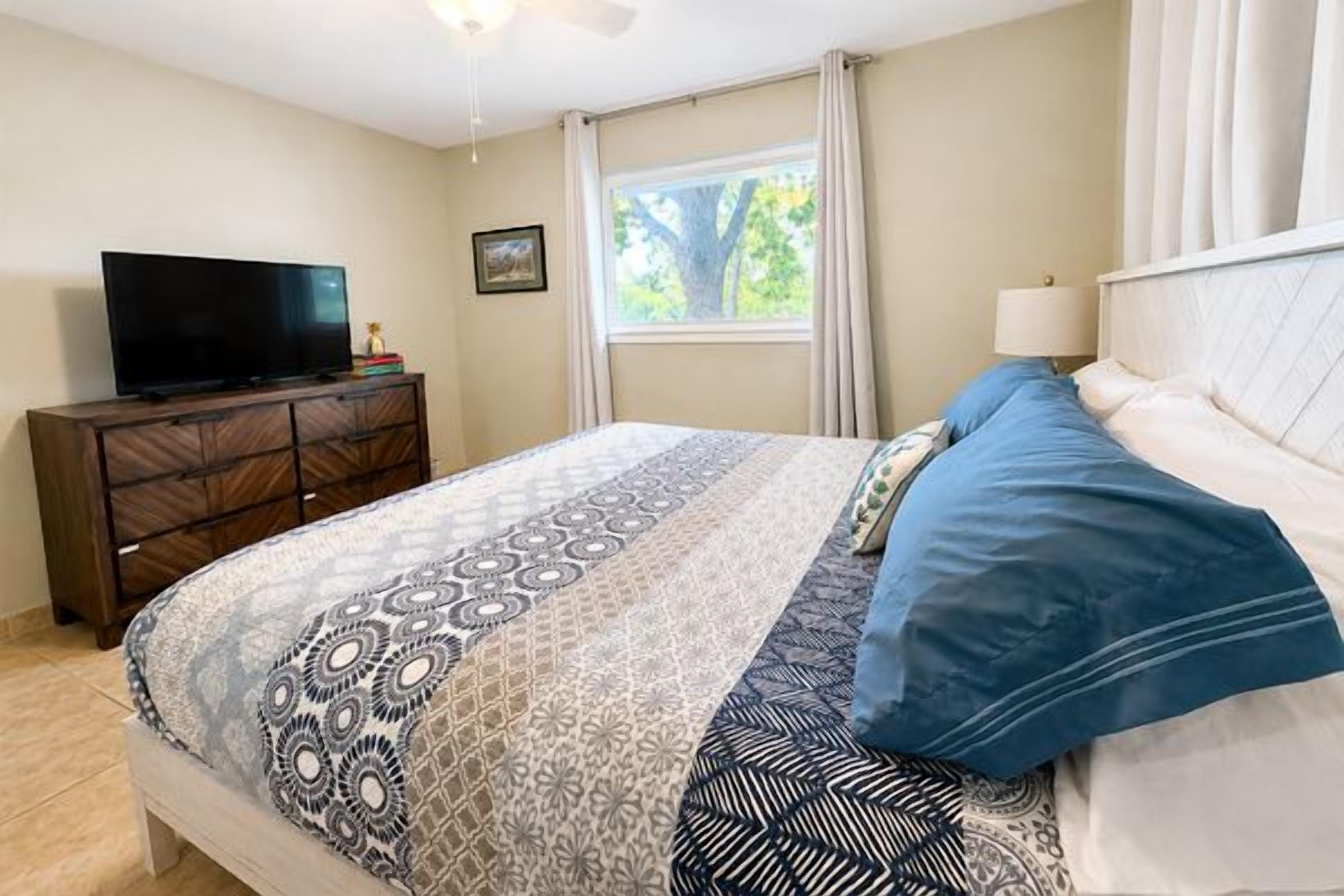 A bedroom with a bed, a television on a dresser, a window with curtains, and a ceiling fan.