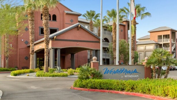 Entrada de un hotel con fachada roja, palmeras y un letrero que dice 'Wyndham by Wyndham'.