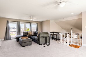 Ein modernes Wohnzimmer mit einem Ecksofa, einem Hocker und einem Glastisch. Eine Treppe mit weißer Geländer führt auf eine erhöhte Ebene.