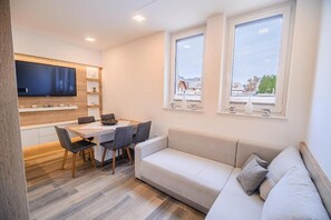 A modern living room with a sofa, a dining table with chairs, a flat-screen TV mounted on the wall, and a window with a view of the outside. - Apartment Gorski Raj (Kranjska Gora)